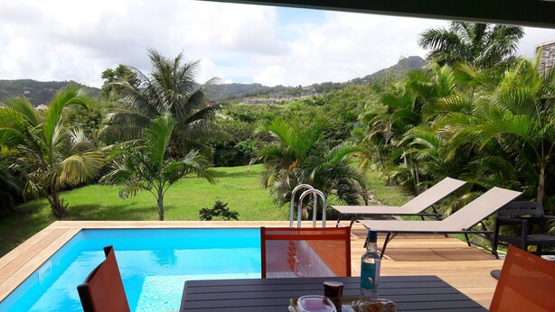 A poolside area with a wooden deck, a table with chairs, and a view of a lush garden and hills in the background. - Colibri bungalow and pool (Vauclin)