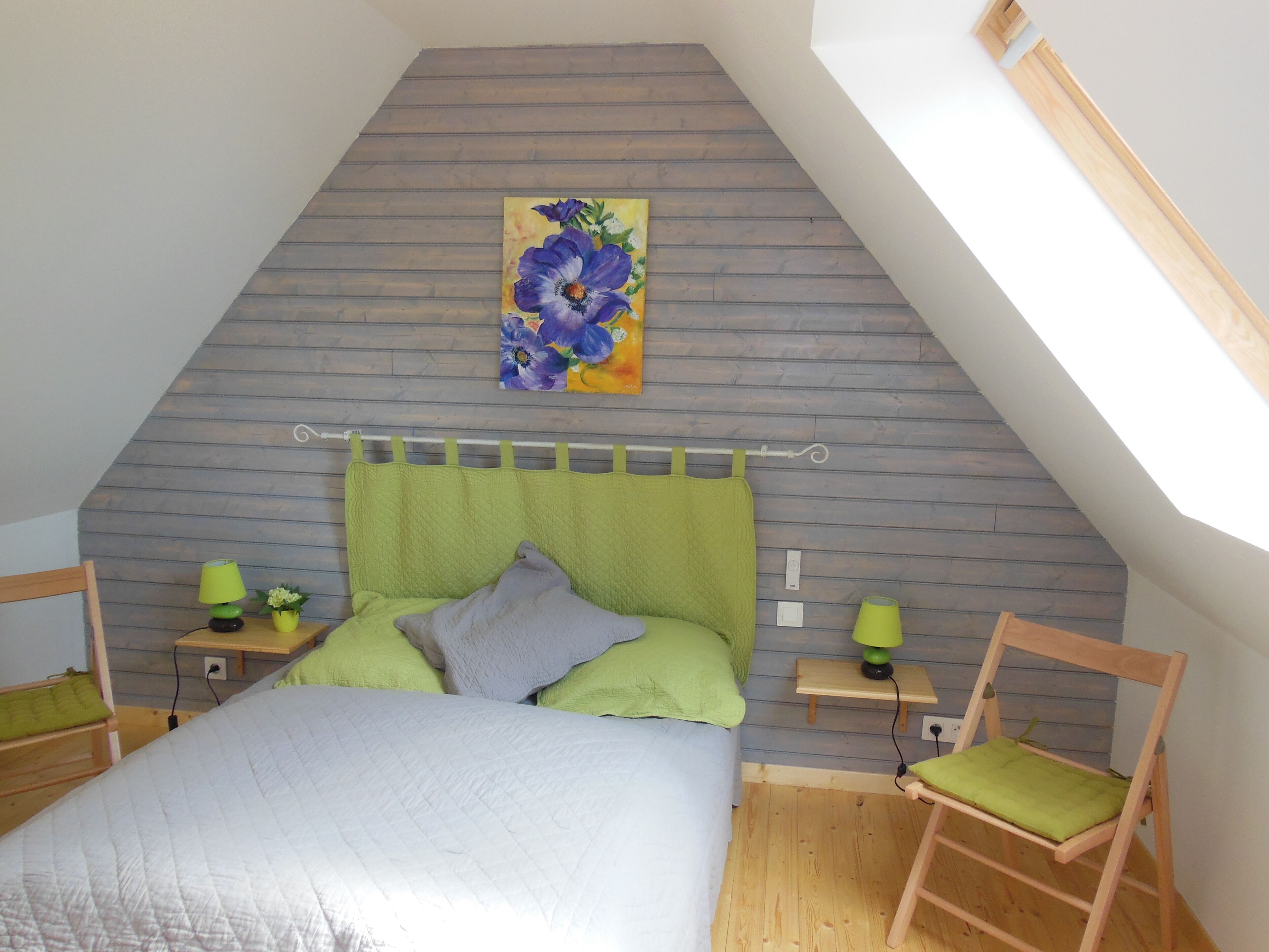 Een slaapkamer met een schuine kap, een bed met een groene hoofdbord, een nachtkastje, een stoel, en een schilderij van bloemen aan de muur.