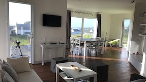 A spacious living room with a sofa, coffee table, and a dining area with a table and chairs. A television is mounted on the wall, and there is a balcony with a tripod.