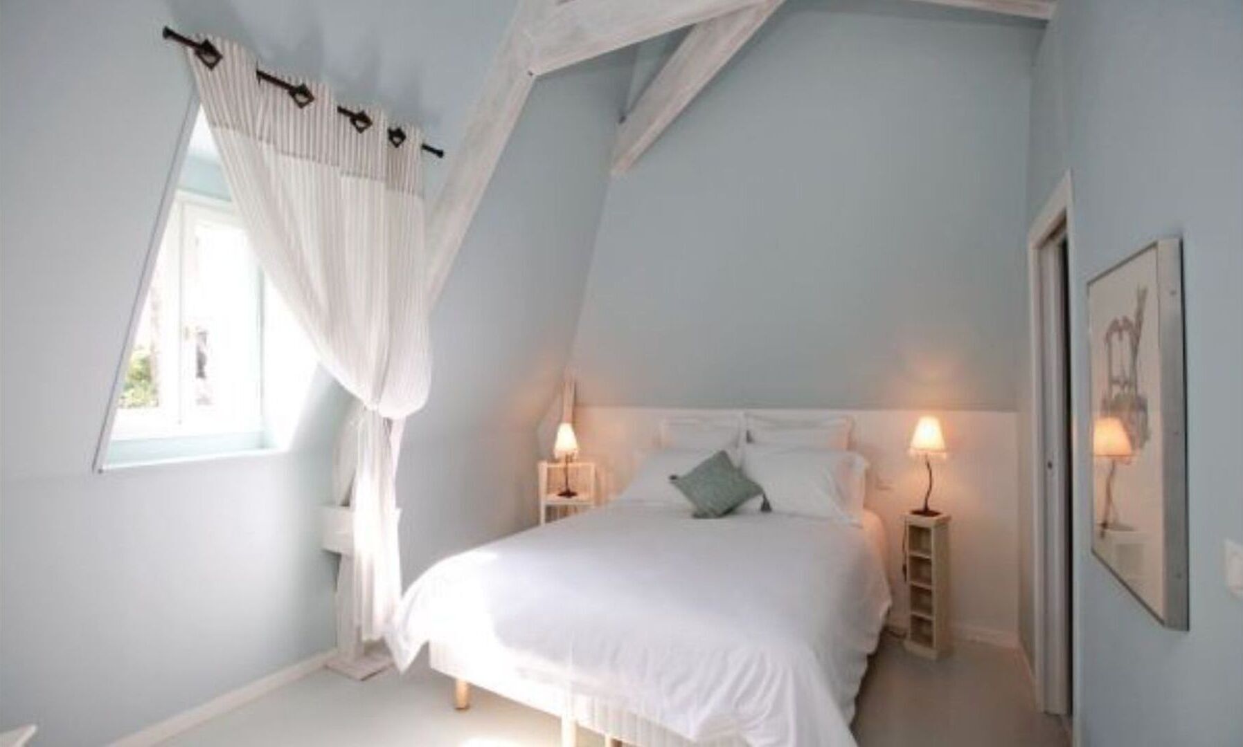 Een slaapkamer met een schuine kap, een bed met witte beddengoed, twee bedlampjes en een raam met witte gordijnen.