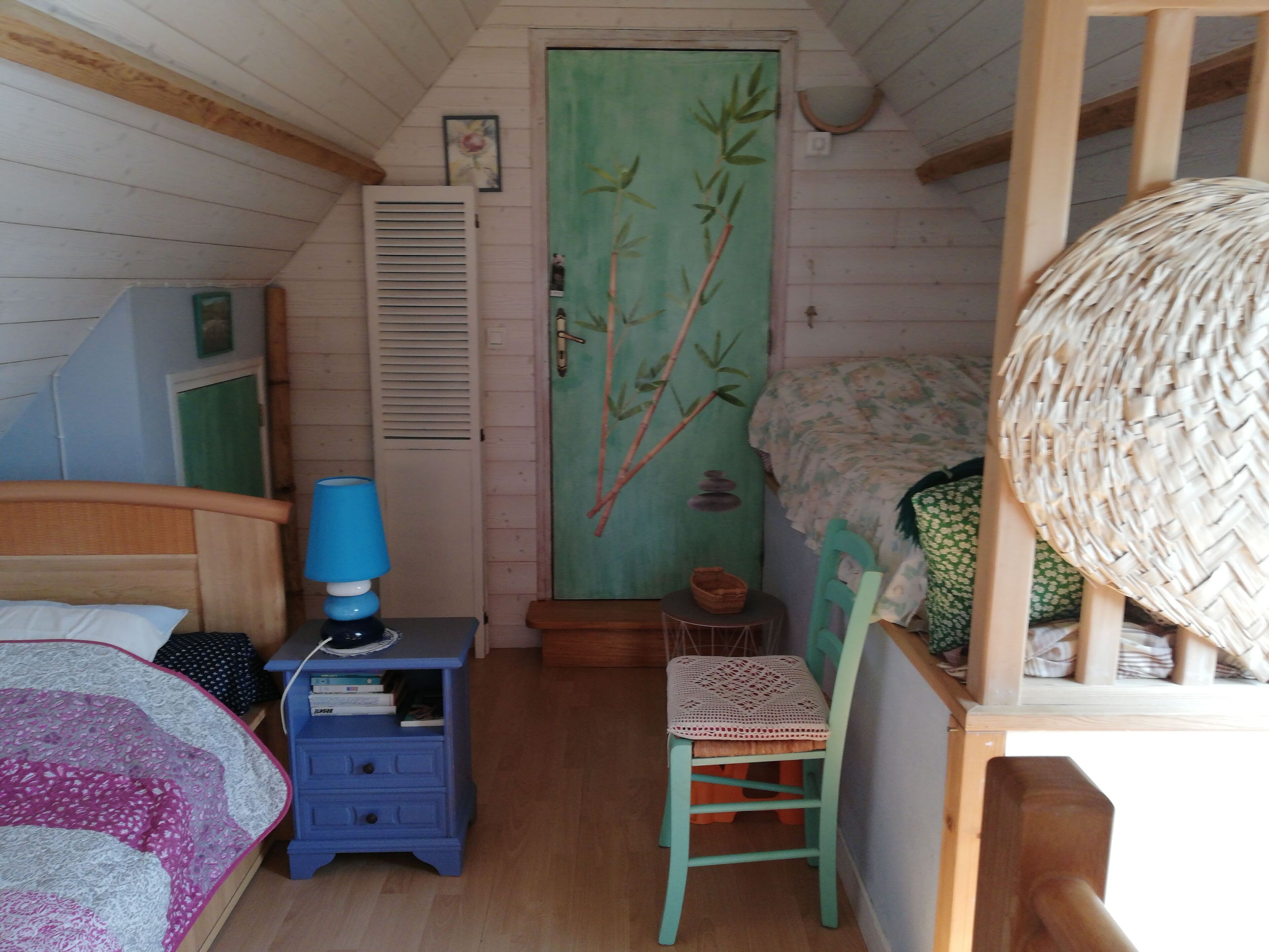 Une chambre confortable avec un lit, une table de chevet bleue, une chaise en osier, une porte verte ornée d’une peinture représentant du bambou et un plafond en bois.