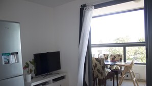 Een kamer met een flatscreen televisie op een witte tv-meubel, een koelkast, en een eethoek met een tafel en stoelen.