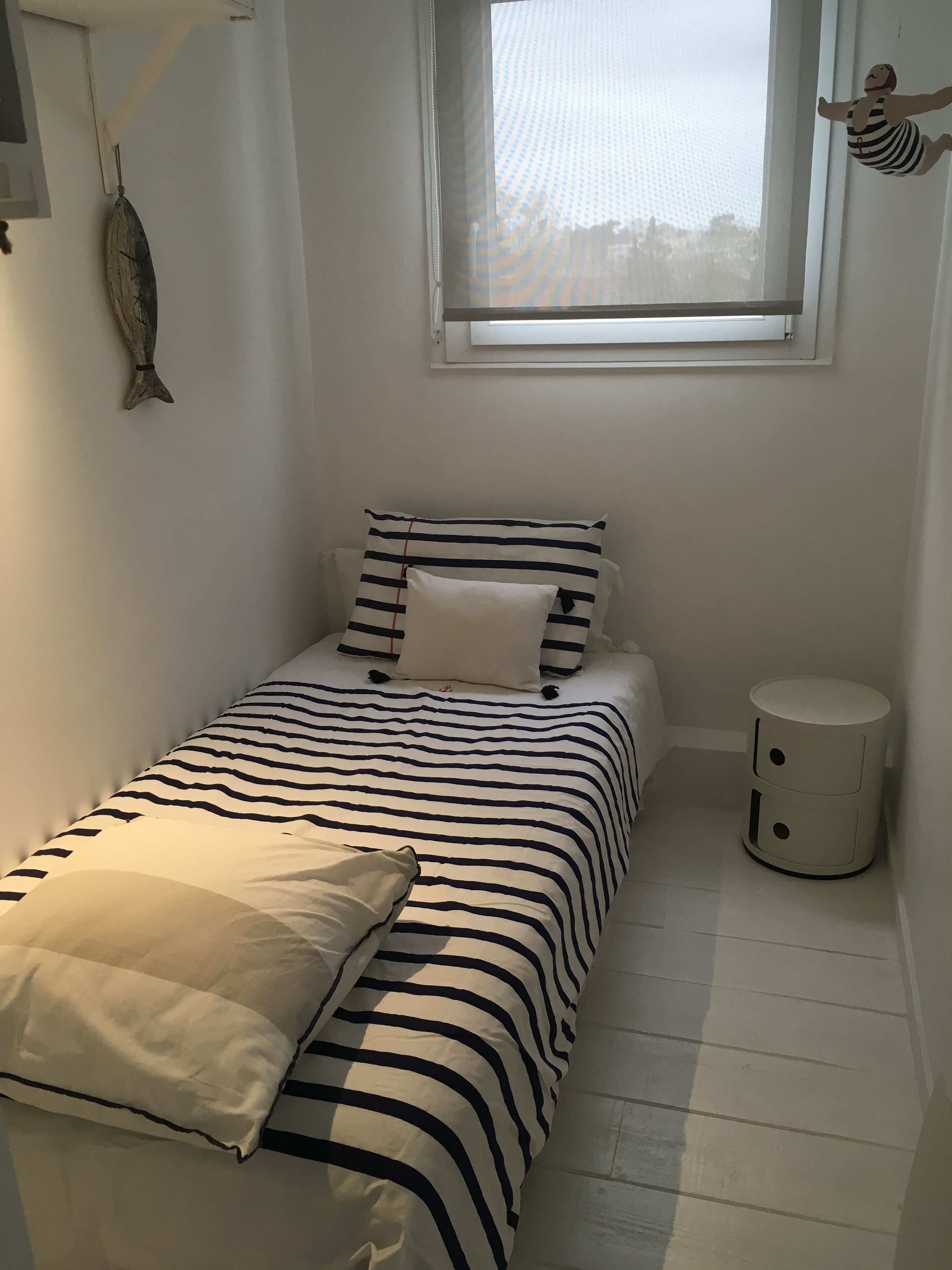 A small, neatly arranged bedroom with a bed featuring striped bedding, a bedside table, a round nightstand, a fish hanging on the wall, and a window with a view of the sky.