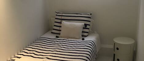 A small, neatly arranged bedroom with a bed featuring striped bedding, a bedside table, a round nightstand, a fish hanging on the wall, and a window with a view of the sky.