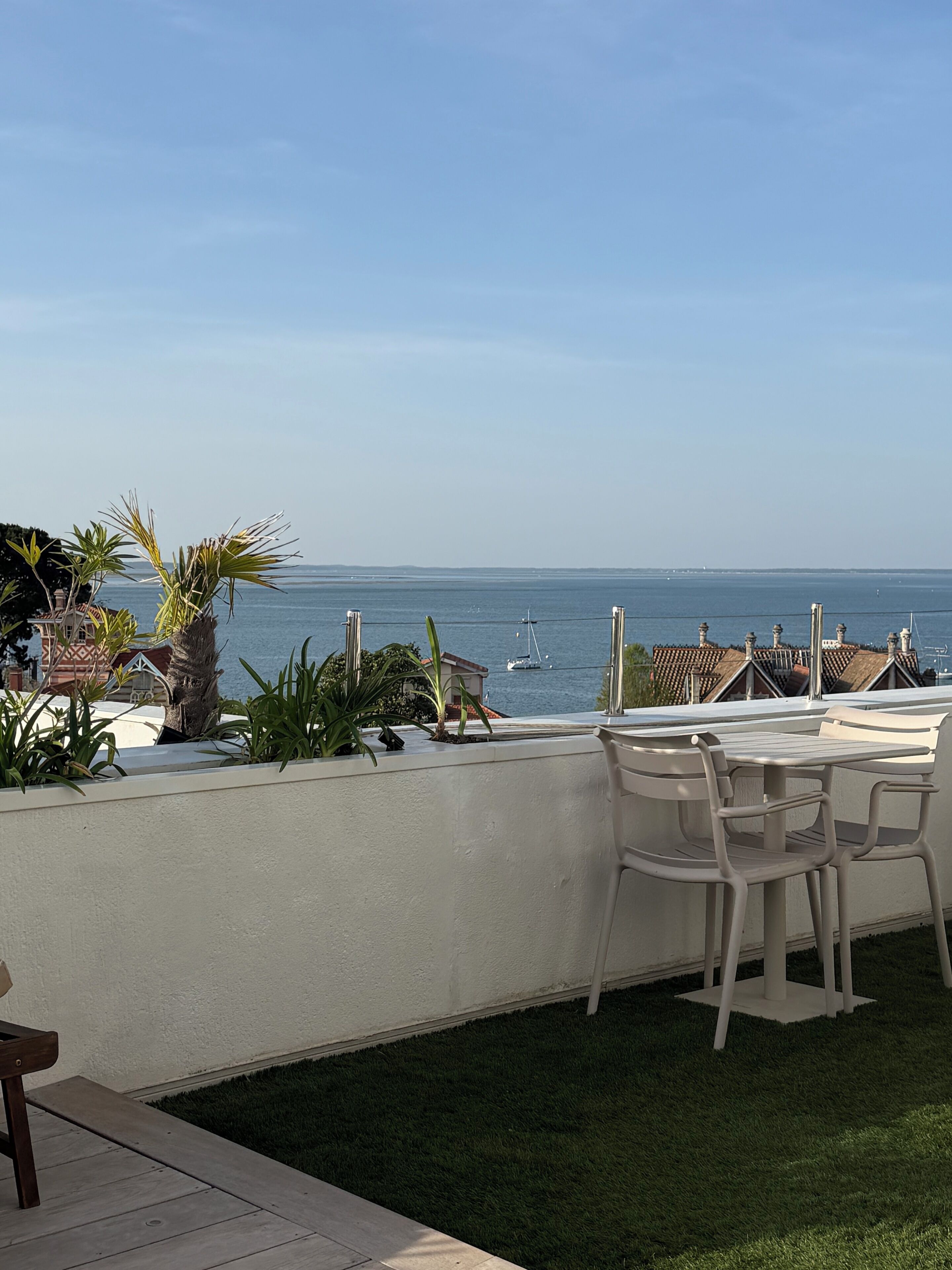 An outdoor patio with a view of the sea, featuring a table and chairs, and a small garden area with plants.