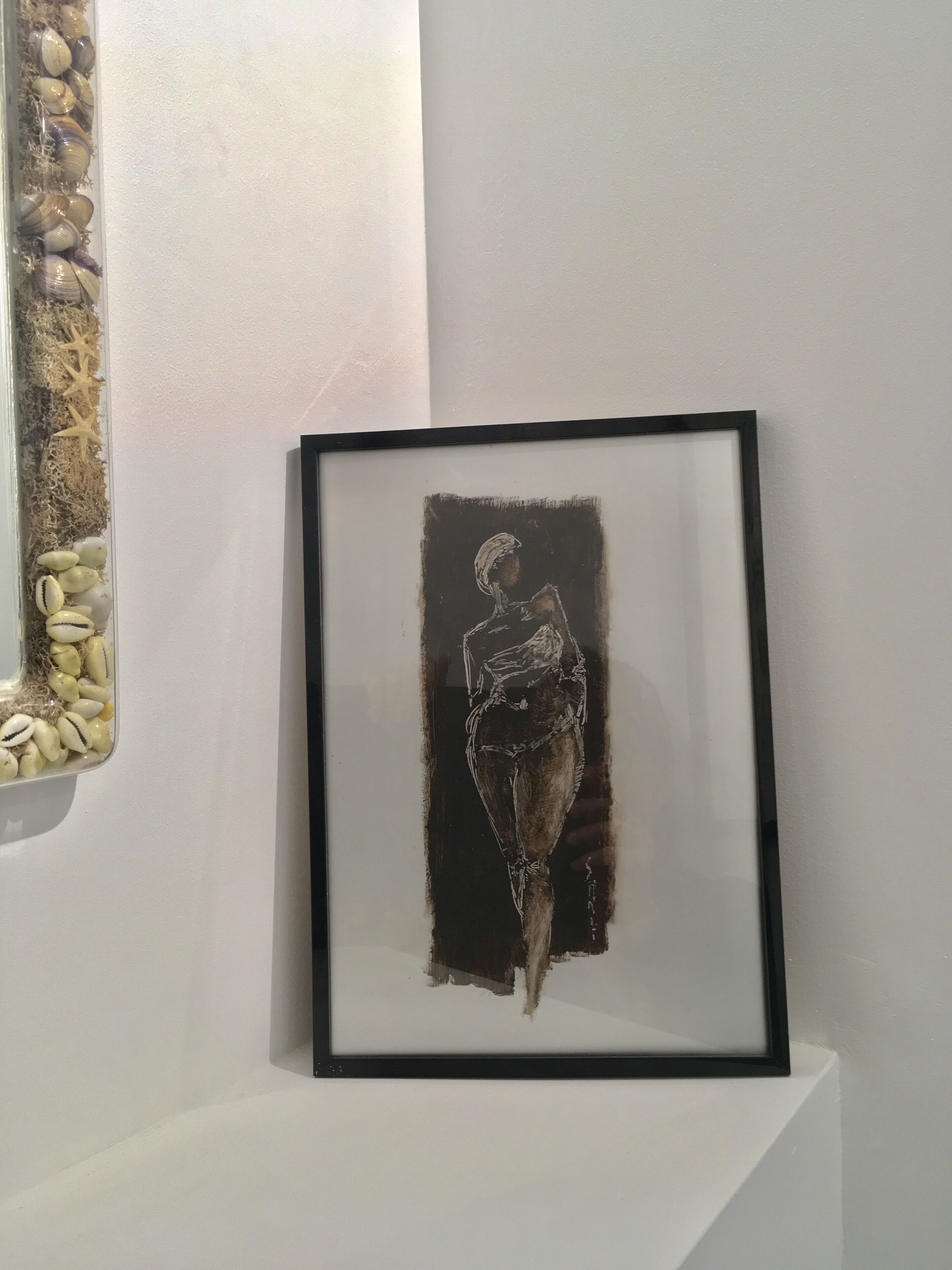 A framed artwork on a white wall, a window with a view of the beach, and a shelf with decorative items.