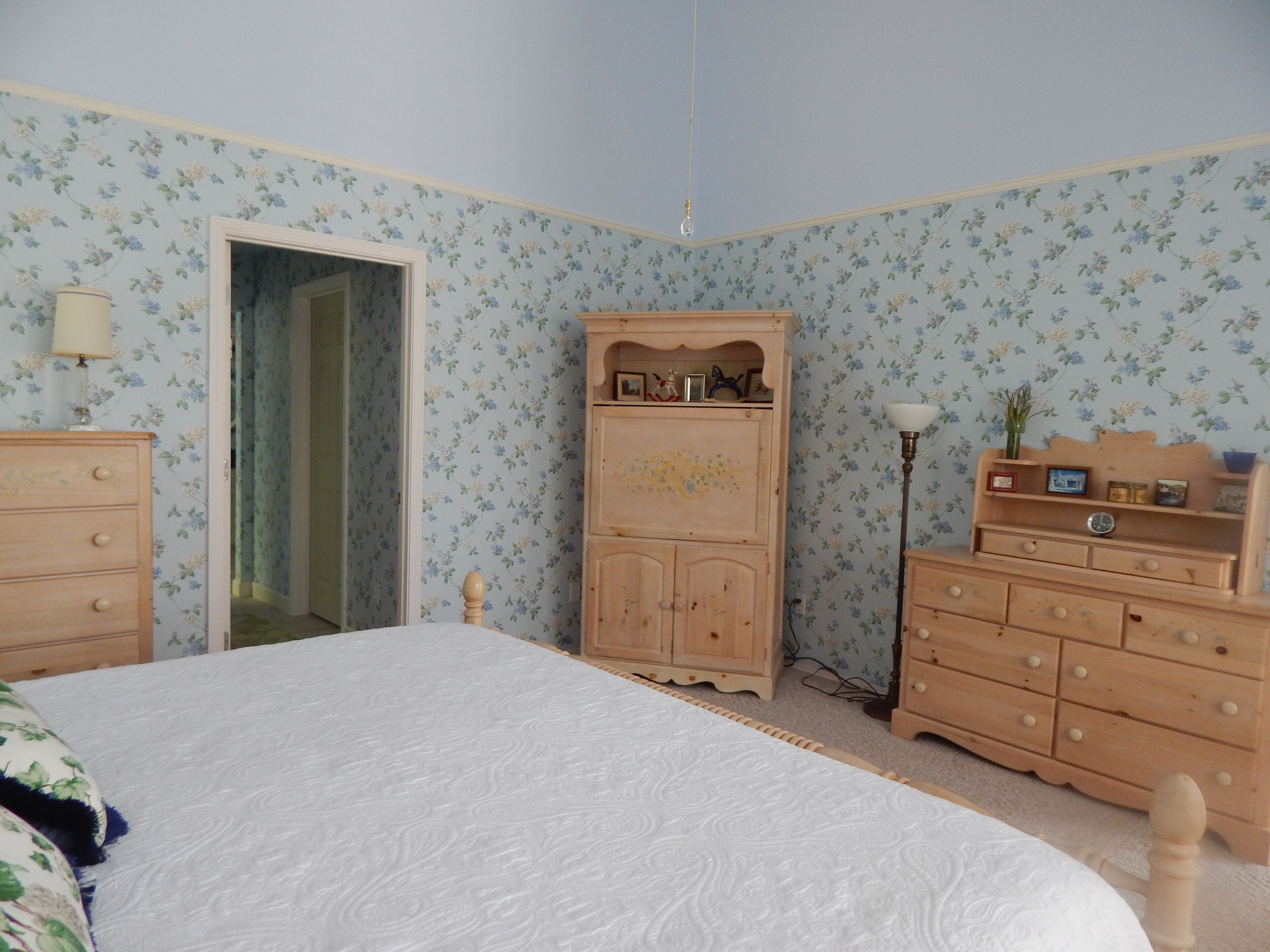 Une chambre avec un papier peint à motifs floraux, une armoire en bois, une table de chevet avec une lampe et un lit recouvert d’une couvre-lit à motifs.
