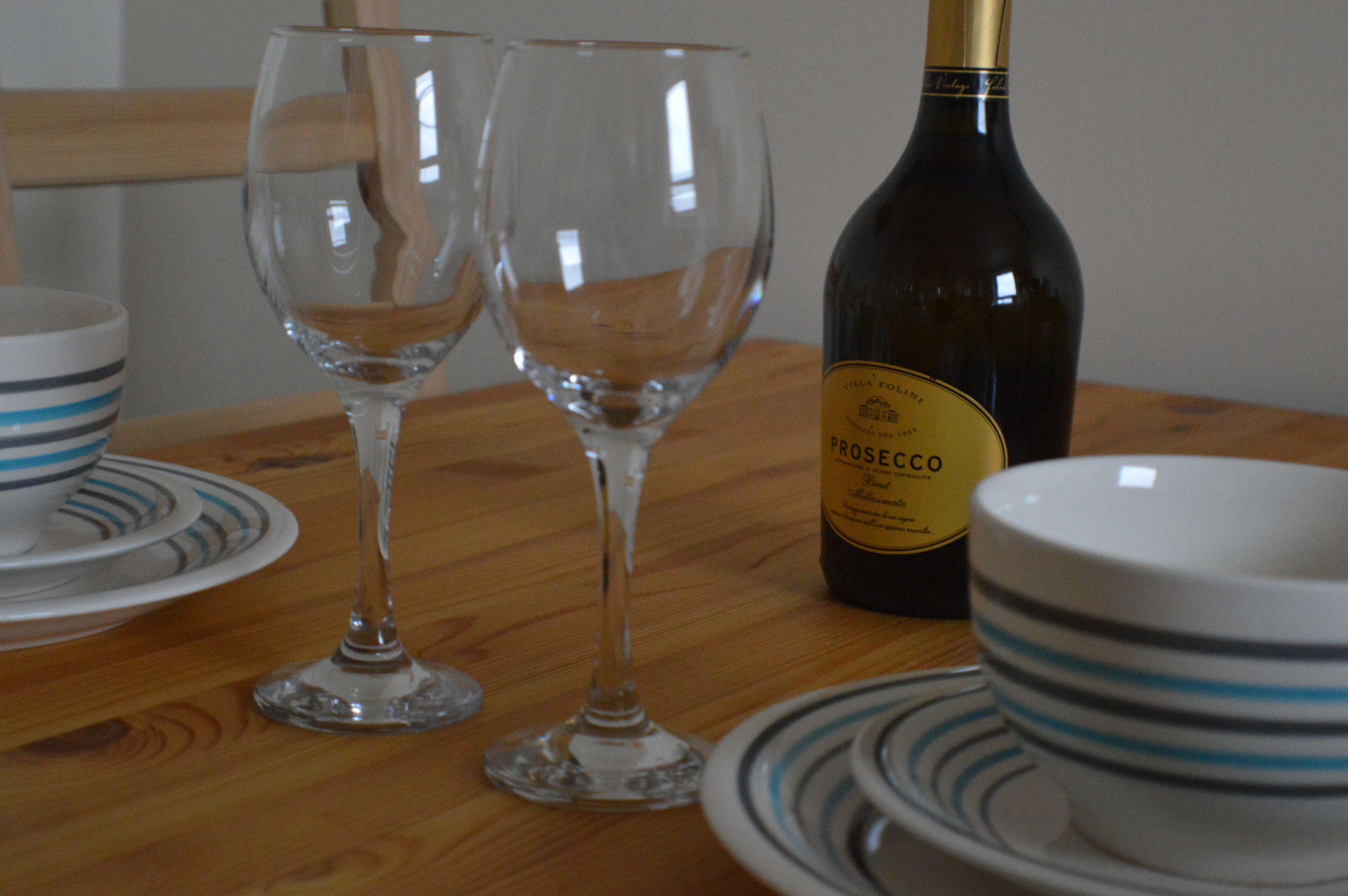 A wooden table with two empty wine glasses, a bottle of Rosecco wine, and striped bowls.