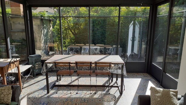 Une pièce baignée de lumière, avec une table et des chaises, des carreaux de sol à motifs et de grandes fenêtres donnant sur un paysage verdoyant.