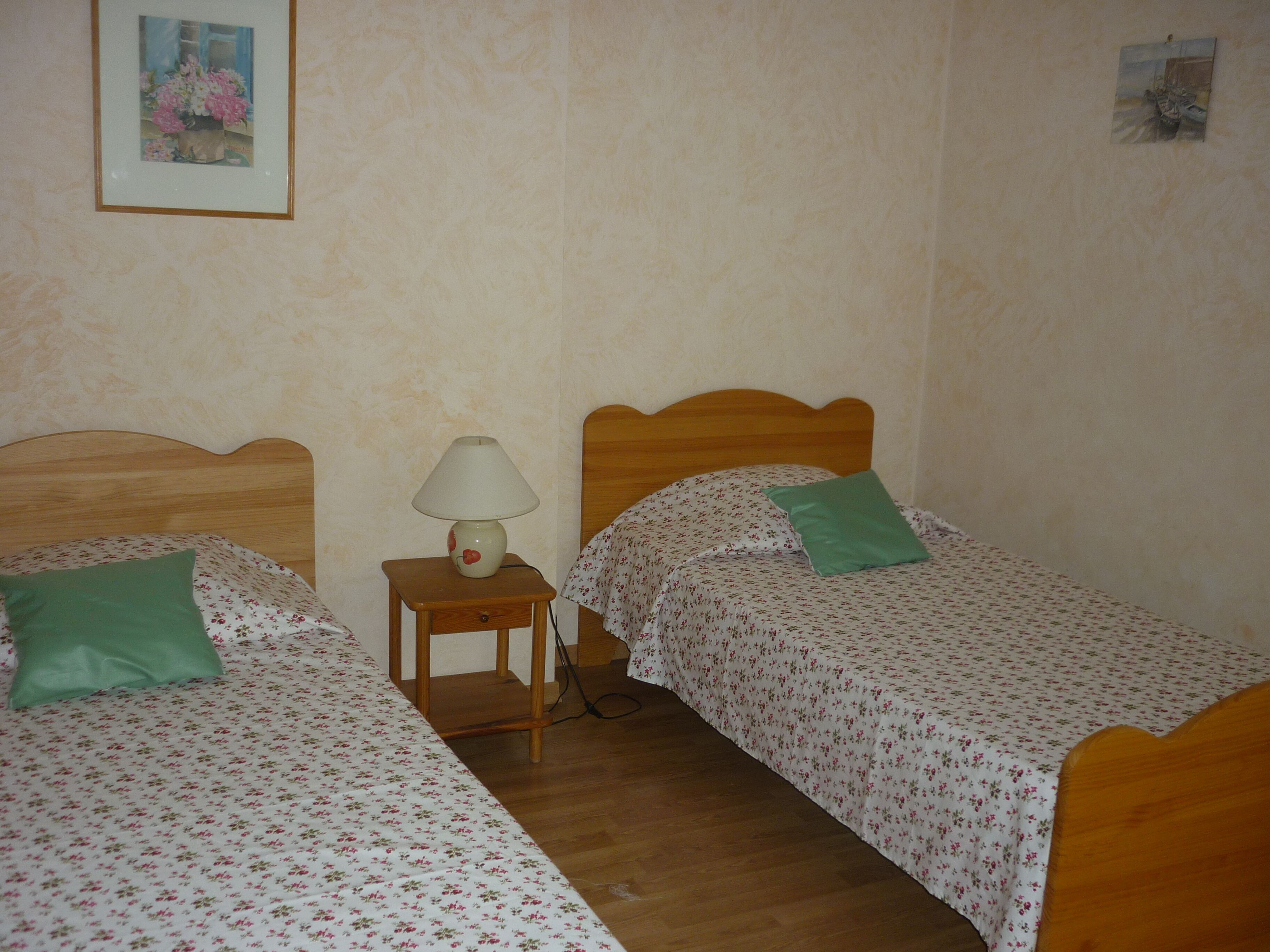 Une chambre avec deux lits simples, chacun recouvert d’une couvre-lit à motifs floraux et agrémenté d’un oreiller vert. Une table de chevet en bois avec une lampe se trouve entre les deux lits.
