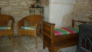 Une pièce avec un mur en pierre, un lit en bois recouvert d’une couverture colorée, deux fauteuils en osier avec des coussins, une table avec une lampe et un poêle d’époque.