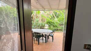 Uma porta que dá para um pátio externo com uma mesa e cadeiras, piso de azulejos e um tapete verde na entrada.