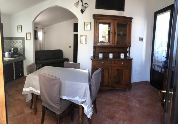 A dining area with a table set for two, a dark sofa, a wooden cabinet with decorative items, and a kitchen area with a coffee machine. - House Delia e Daniela (Terrasini, Palermo)