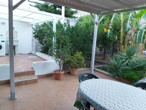 A covered patio area with a table and chairs, surrounded by potted plants and a small garden. - House Delia e Daniela (Terrasini, Palermo)