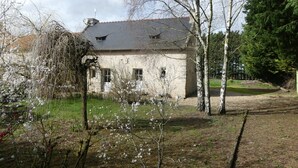 Une maison de campagne au toit en pente, entourée d’arbres et bordée d’une cour gazonnée.
