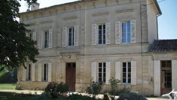 A two-story building with a symmetrical facade, featuring evenly spaced windows with shutters, a central entrance, and a small garden area with a chair and plants.
