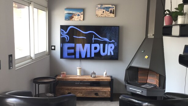 A living room with a TV displaying 'EMPUR', a wooden entertainment unit, a black grill, and two black leather sofas. - House for rent gulf of St Tropez 2 bedrooms in residence with swimming pool tennis (Gassin)