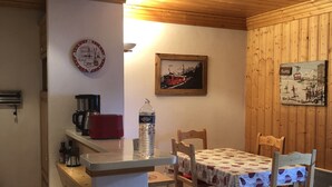 Um quarto com painéis de madeira, uma mesa de jantar posta para quatro pessoas, uma cafeteira, uma garrafa de água e um relógio na parede.