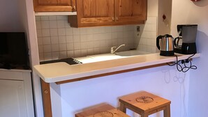 Uma cozinha com armários de madeira, bancada branca, pia, cafeteira e dois banquinhos de madeira.