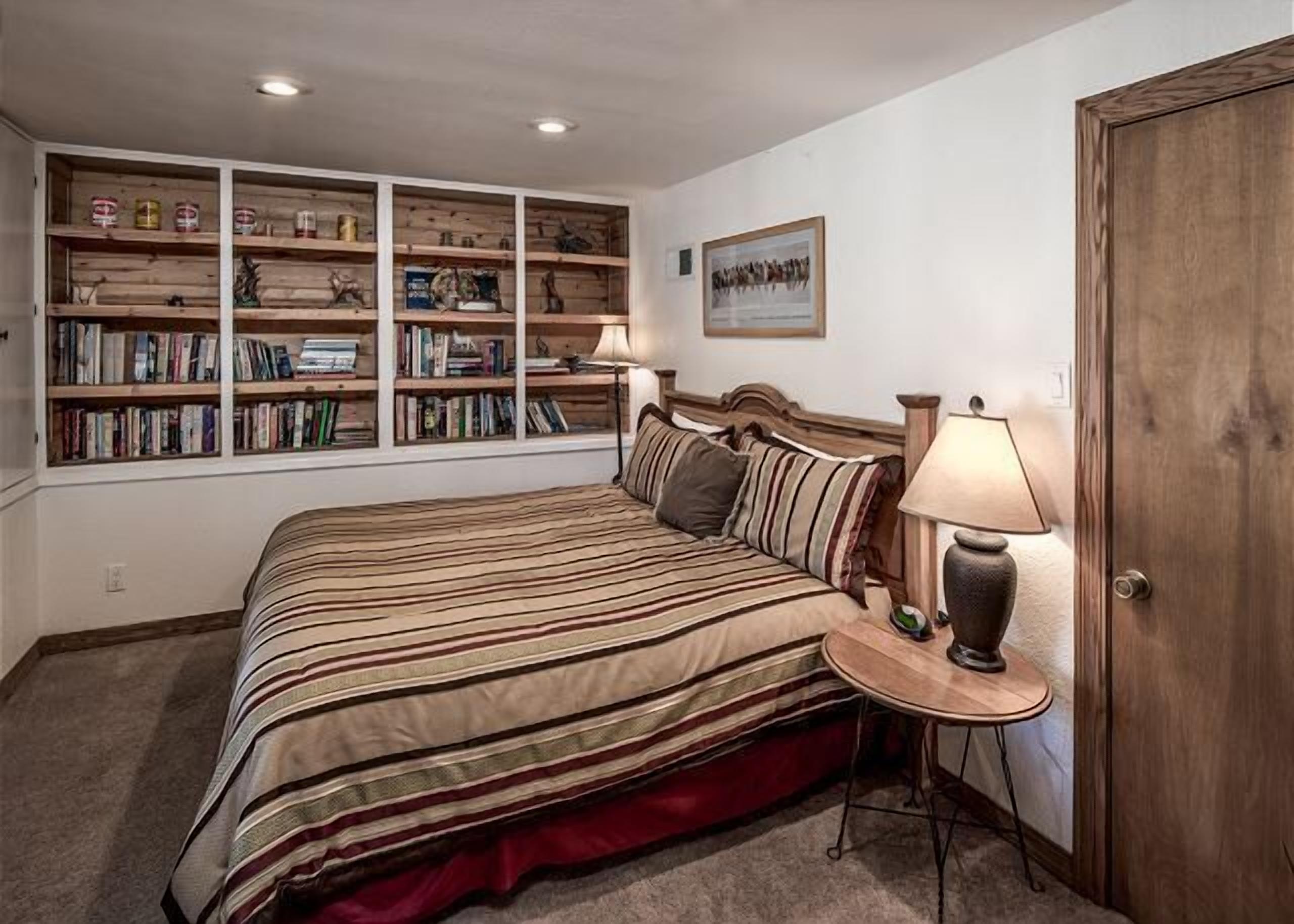 A bedroom with a large bed, a bedside table with a lamp, a wooden bookshelf, and a door.