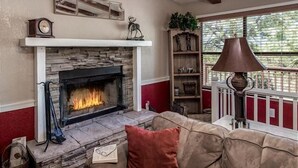 A cozy living room with a stone fireplace, a red and beige sofa, a bookshelf, and a window with a view of trees.