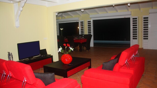 A modern living room with red sofas, a black coffee table, and a TV. There is a dining area with a table and chairs visible through an open door.
