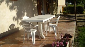 Um pátio com uma mesa e cadeiras brancas, rodeado por um jardim e um edifício com telhado em cerâmica.