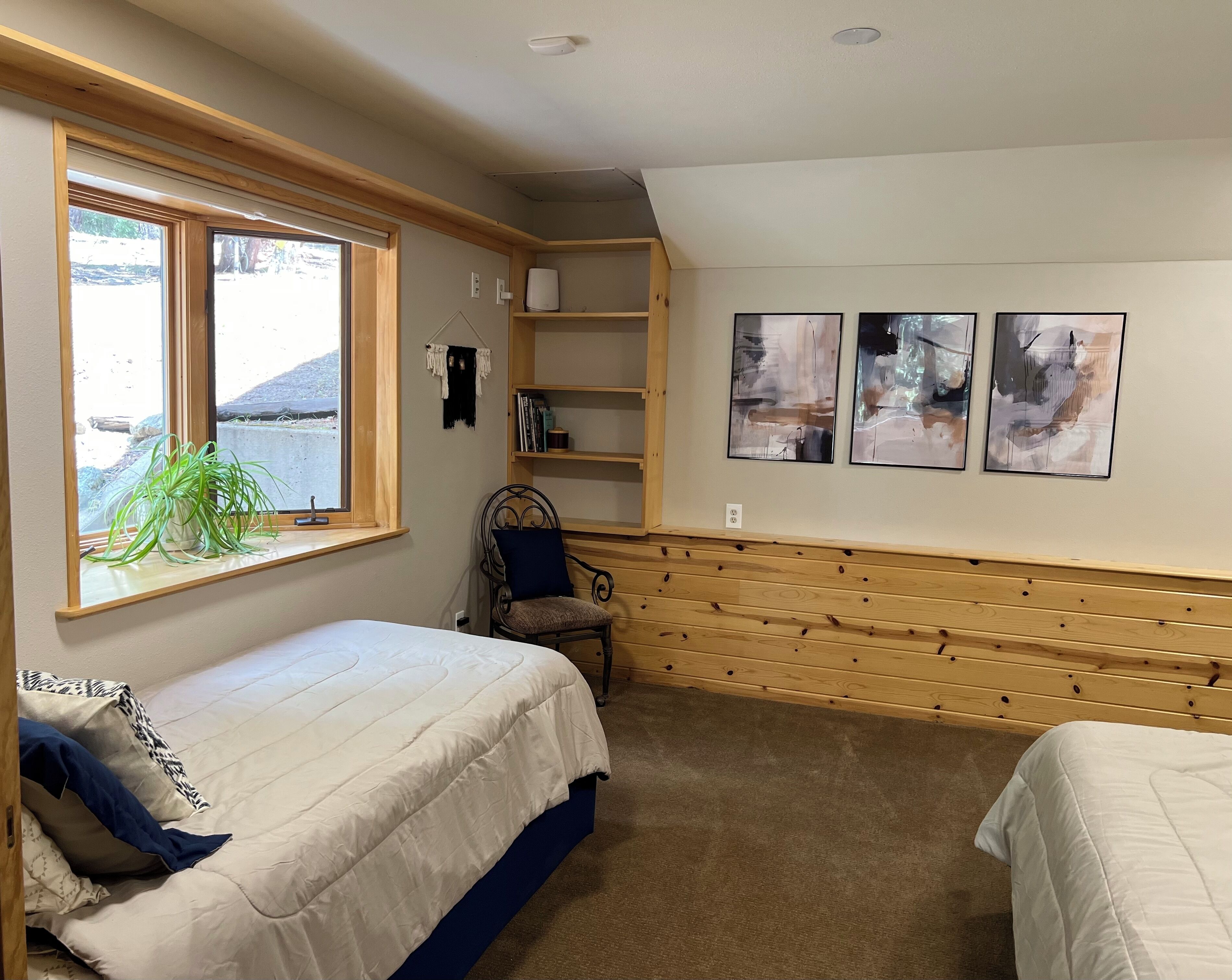 Un dormitorio con dos camas, una ventana con vistas, una silla, una estantería con libros y arte abstracto en la pared.