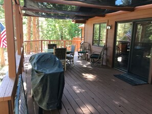 A covered wooden deck with outdoor furniture, including a covered grill and chairs, and a sliding glass door to the house. - Red Fox Property Graeagle/Blairsden (Clio)