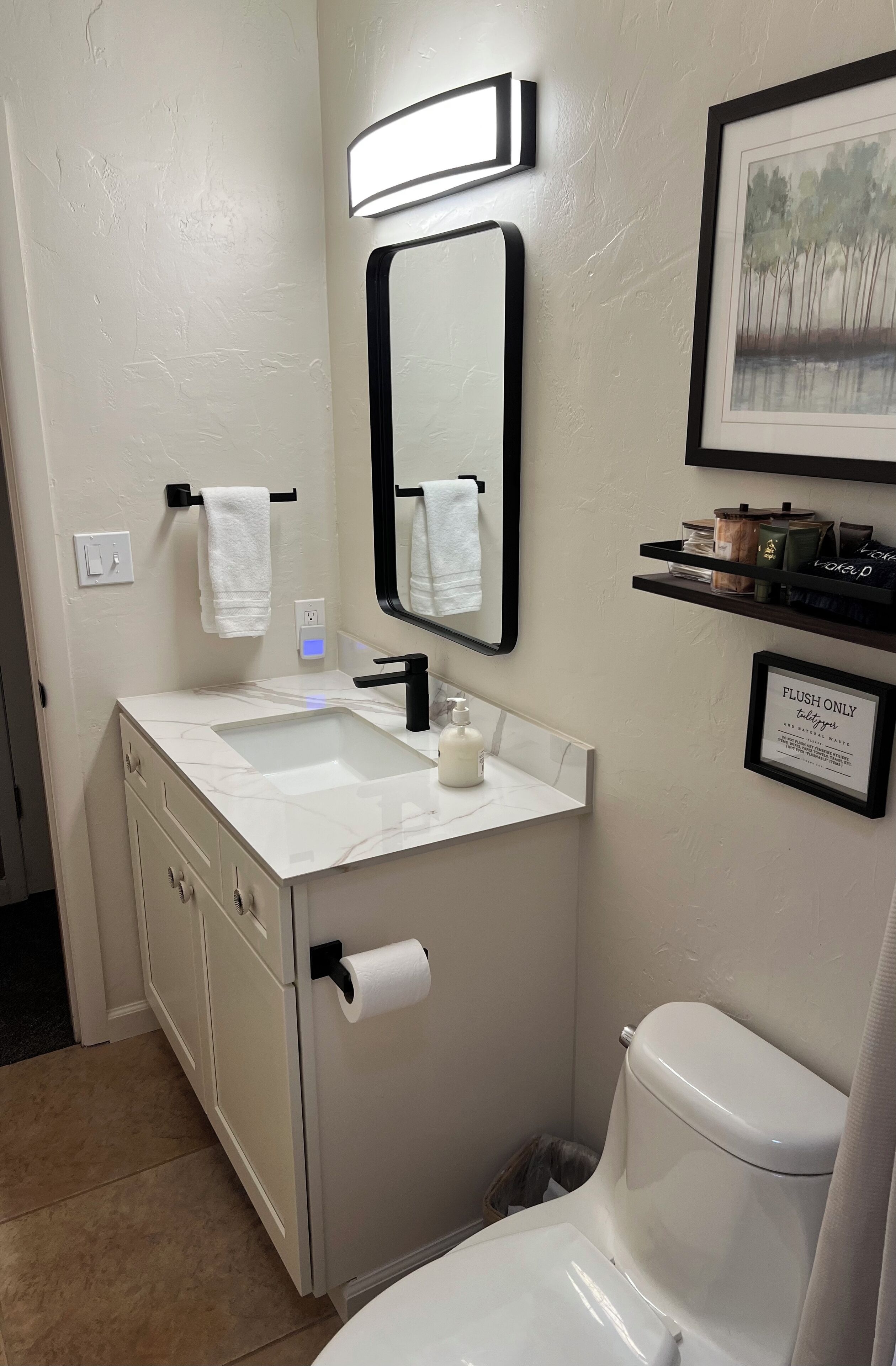 A bathroom with a white sink, black faucet, and a toilet. There is a framed picture on the wall and a shelf with toiletries.