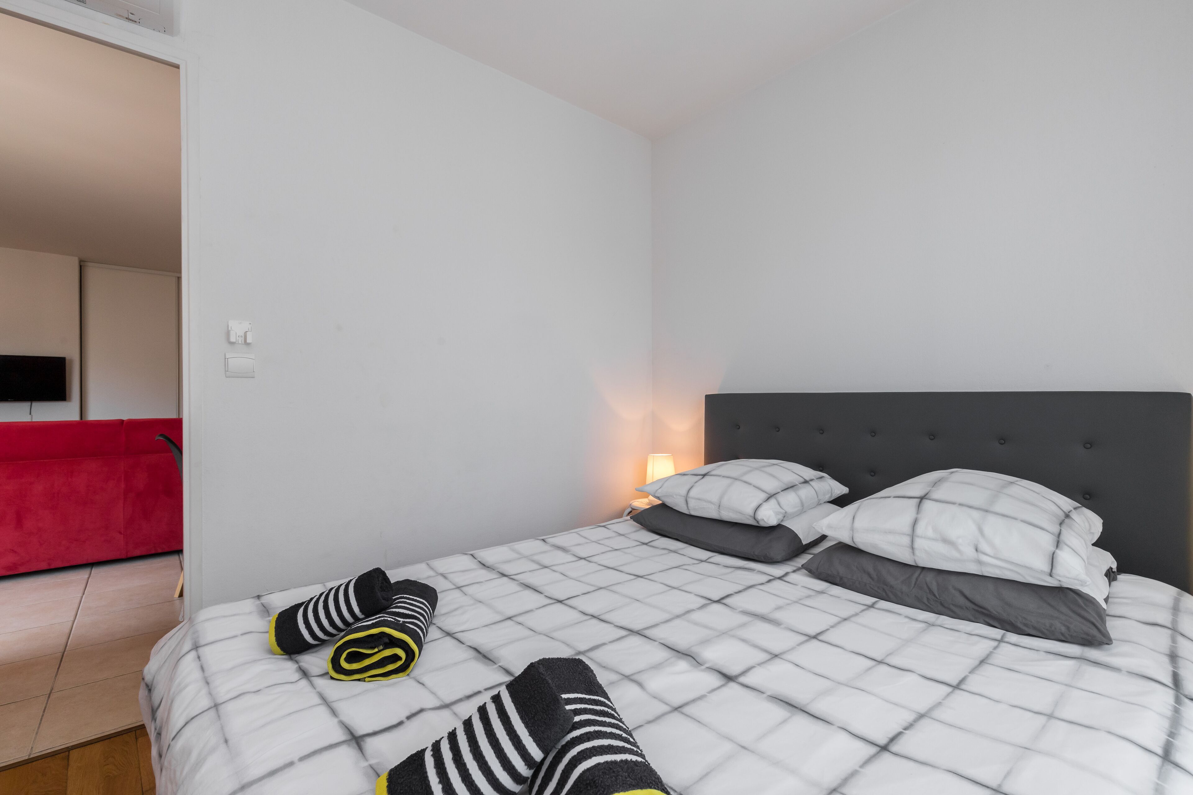 A neatly made bed with striped and solid pillows, a red couch in the background, and a wall-mounted TV.