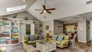 A spacious living room with a high ceiling, a ceiling fan, and a large window. There is a green sofa set, a coffee table, and a bookshelf filled with various items.