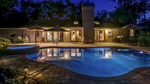 A swimming pool with a patio area, outdoor seating, and a building with a chimney in the background.