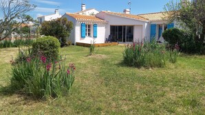 A white house with blue shutters and a tiled roof, surrounded by a well-maintained garden with various plants and flowers. - Very quiet location, 5 minutes from the beach/10 minutes from shops by bike, large garden with castle view/Wifi. (Noirmoutier-En-L'île)