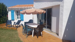An outdoor patio with a table and chairs, a striped umbrella, and a white building with blue shutters. - Very quiet location, 5 minutes from the beach/10 minutes from shops by bike, large garden with castle view/Wifi. (Noirmoutier-En-L'île)
