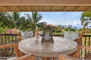 A round table with chairs, a bottle of champagne, and a glass on a covered patio with a view of a house and palm trees.