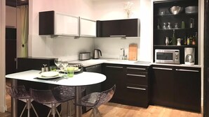 A modern kitchen with dark cabinetry, a white countertop, and a round dining table with transparent chairs. - Charming studio Ile Saint Louis PARIS 4 ° ° - Paris (Paris)