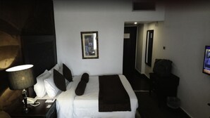 A hotel room with a bed, a nightstand, a lamp, a framed picture on the wall, and a door leading to another room. - Sunfort hotel (Lahore)