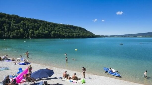 Ein Strand mit badenden und sonnenden Personen, einem klaren Himmel und einer bewaldeten Hügelkette im Hintergrund.