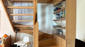 A wooden staircase leading to a loft bedroom with a white chair, a small table, and a TV stand.