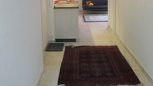Un couloir étroit, avec un tapis marron foncé, une porte blanche et, au fond, on aperçoit un salon avec un canapé et un tapis.