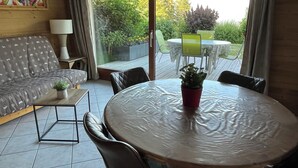 Ein Zimmer mit einer Couch, einem kleinen Tisch mit Pflanze, einem runden Tisch mit Stühlen und Blick auf einen Garten durch eine Glastür.