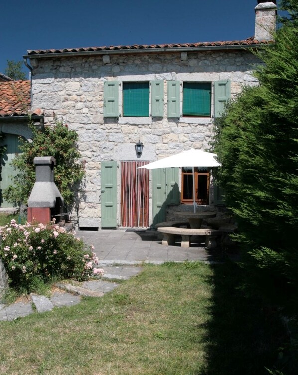 A stone building with green shutters, a stone chimney, and a small garden with a bench.