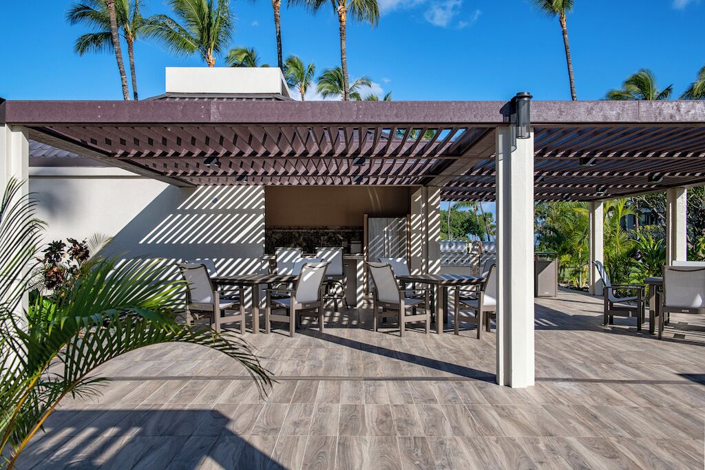 An outdoor patio with tables and chairs, surrounded by palm trees and a pergola structure.