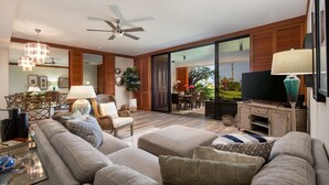 A modern living room with a large sofa, a television, and a dining area visible through glass doors. - Prime Location, Unit H-102, Oceanfront next to pool. (Kamuela)