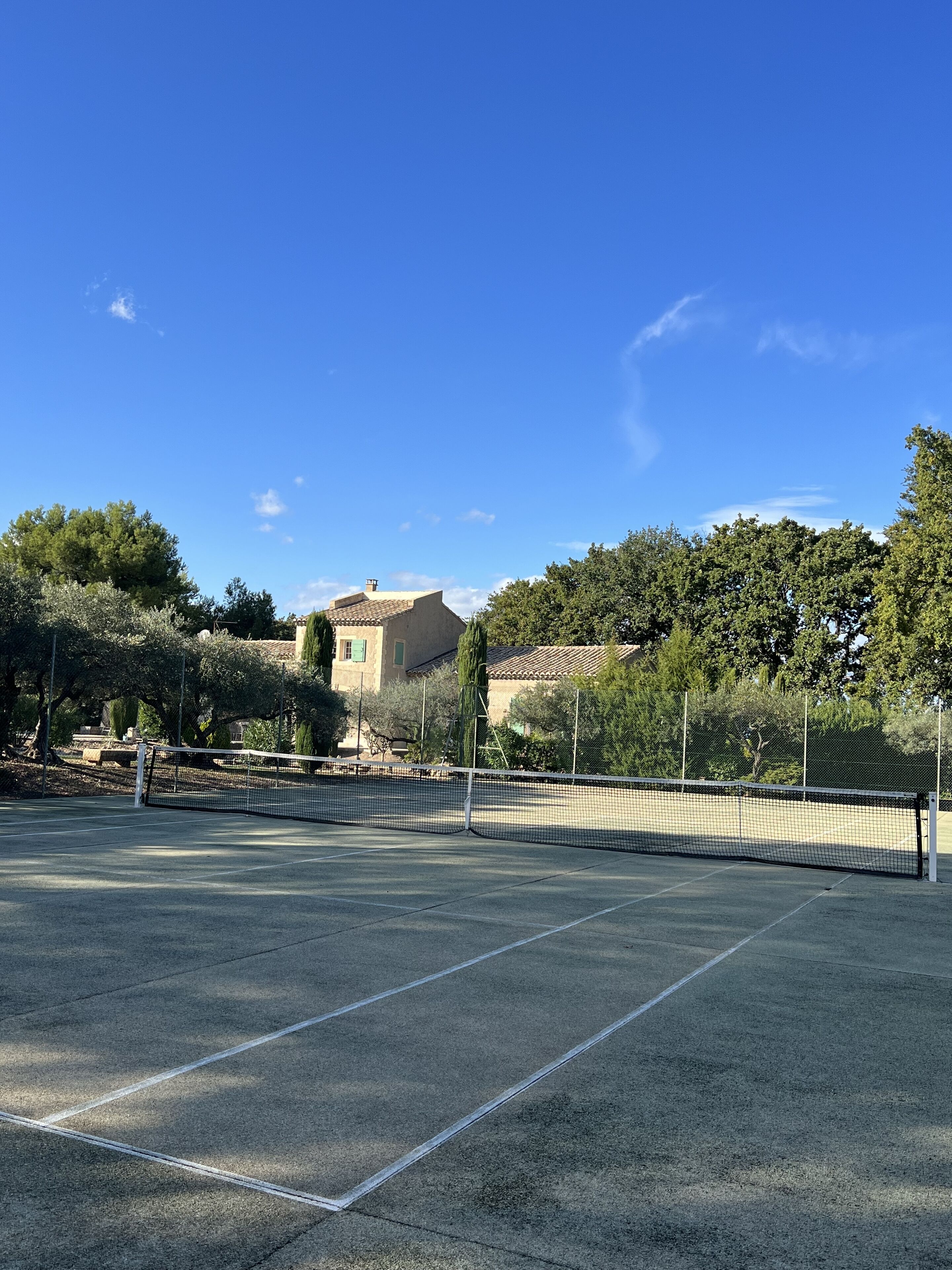 An outdoor tennis court with a net, surrounded by trees and a building in the background.