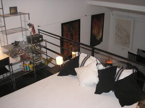Una habitación de hotel con una cama, un escritorio con lámpara, estantería con objetos varios y obras de arte en las paredes.