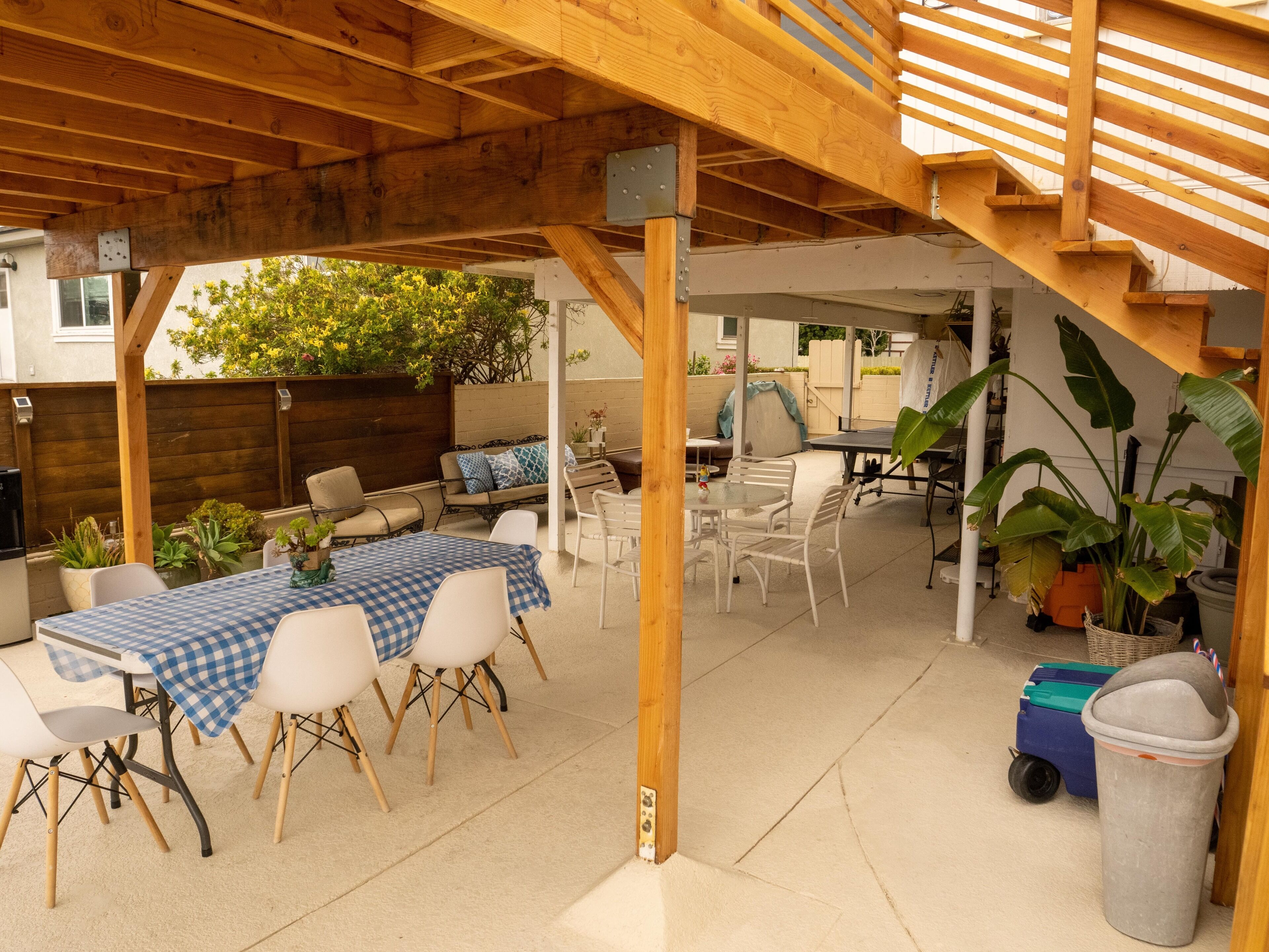 Un espace extérieur aménagé en terrasse, avec une pergola en bois, une table à carreaux, des chaises blanches et des plantes en pot.
