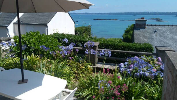 Un patio con una mesa y una silla blancas, rodeado de exuberante follaje y flores, con vista a un cuerpo de agua con barcos.