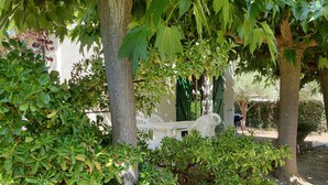 Een overdekt terras met een witte tafel en stoelen, omgeven door weelderige begroeiing en bomen.